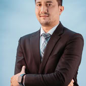A confident businessman posing with arms crossed wearing a formal suit against a blue background.